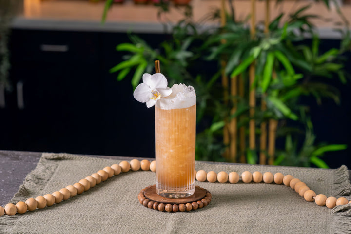 A frothy orange cocktail in a clear highball glass, garnished with a white orchid flower and a small brown pick, resting on a round wooden coaster. The drink sits on a beige textured cloth with a string of wooden beads draped around it, with a blurred green plant and dark background behind.