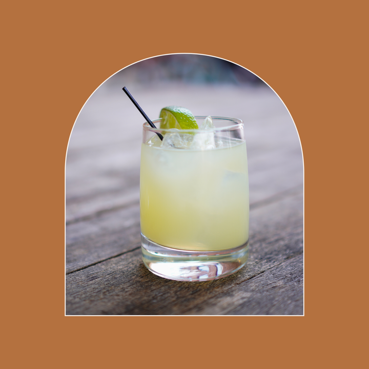 Cocktail with lime wedge and black straw in a glass on a wooden surface, framed by an arch on a brown background