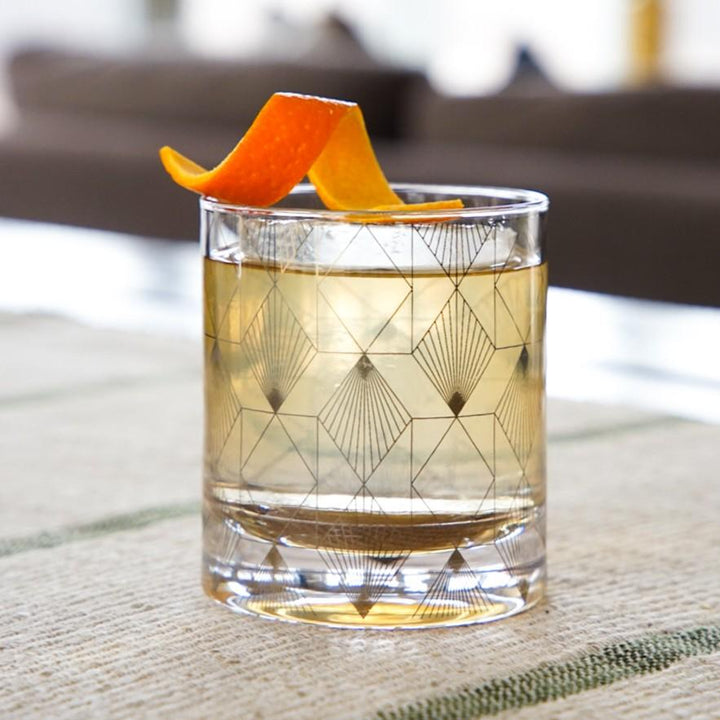 Cocktail in a geometric-patterned glass with orange peel garnish on a blurred background.