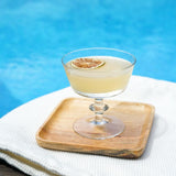 Cocktail in a glass with a lemon wheel on a wooden coaster by a pool.