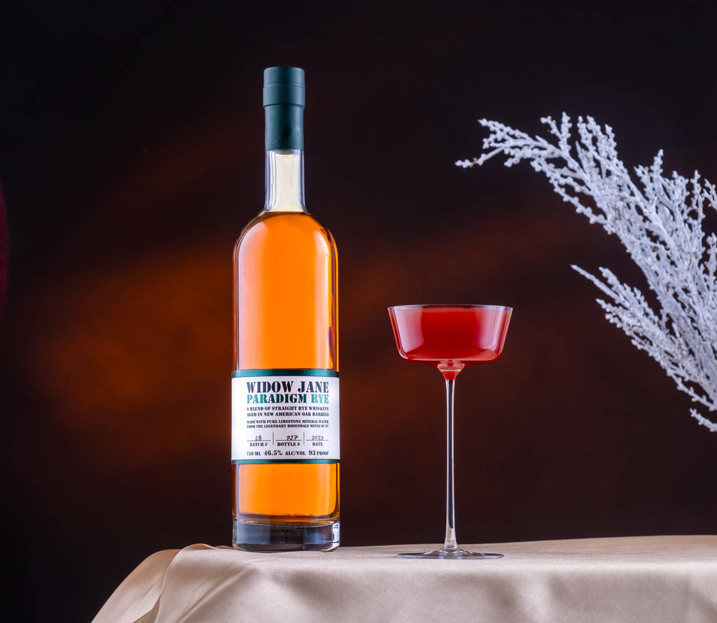 Bottle of Widow Jane Paradigm Rye on a table next to a bright red cocktail.