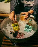 Two people holding glasses of a yellow liquid next to bottles on a tray of ice.