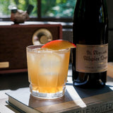 Cocktail with ice and a slice of fruit next to a bottle of St. Elizabeth's Allspice Dram on a book outdoors.