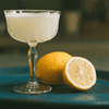 Image of chilled cocktail in a coup next to a lemon garnish.