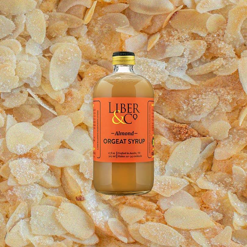 Bottle of Liber & Co. Almond Orgeat Syrup over backdrop of sliced almonds.