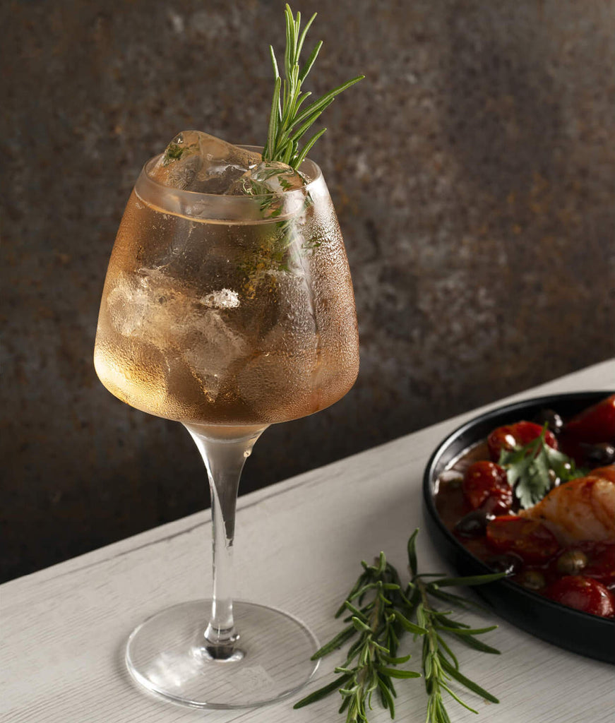 Cocktail garnished with rosemary on a table next to a plate of food.