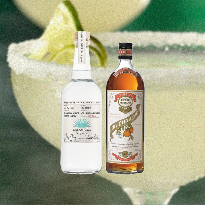 Bottles of Casamigos Blanco Tequila and Pierre Ferrand Dry Curacao. over close up of a margarita in a glass with limes and salted rim.