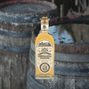 Bottle of Fortaleza Tequila Añejo imposed over an image of two rustic barrels.