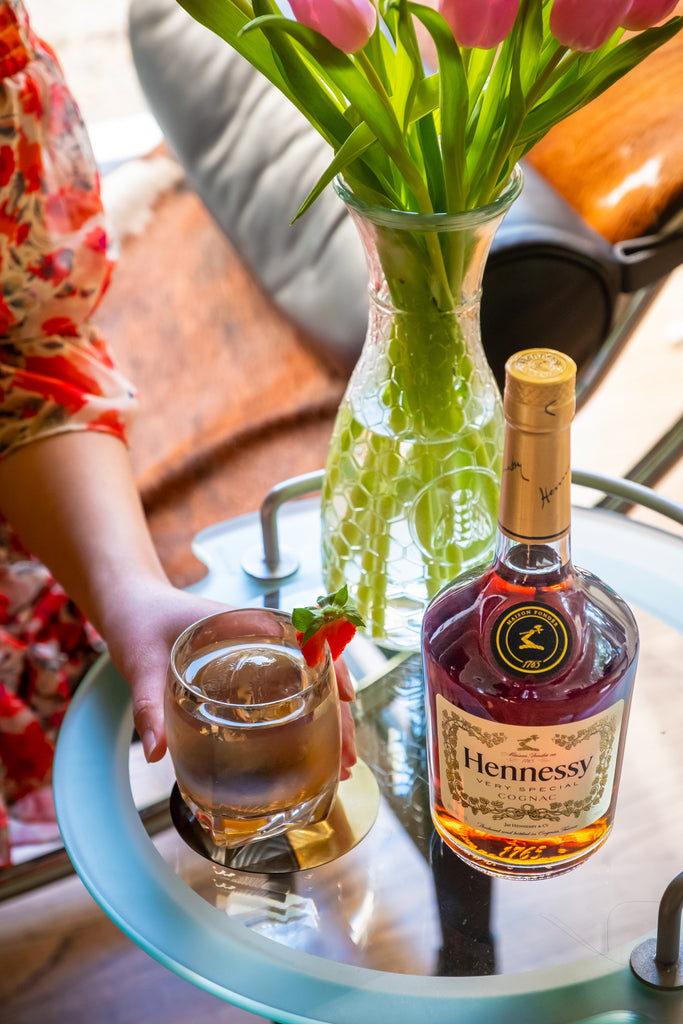 Bottle of Hennessy V.S. Cognac, positioned on a glass table next to a vase of pink tulips, and a craft cocktail garnished with a strawberry.