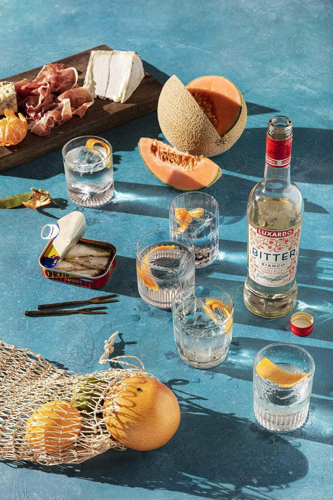 Tablescape of cantaloupe, cocktails, cheese and meat board and sardines.