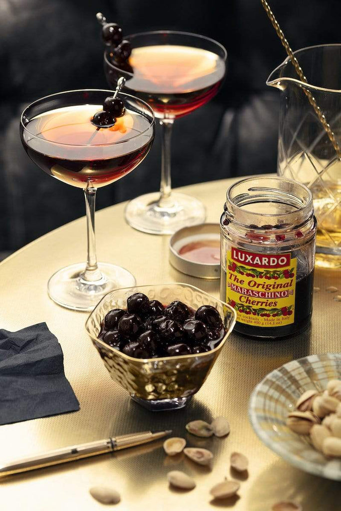 Luxardo Maraschino Cherries Jar on a table with a bowl of cherries and two cocktails.