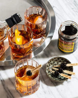 Open jar of Luxardo cherries on a table with three cocktails.