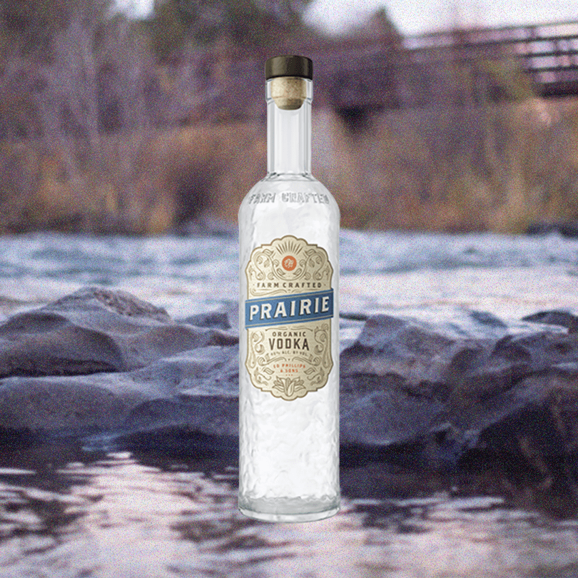 Bottle of Prairie Organic Vodka over backdrop of a shallow stream with an old walking bridge in background.