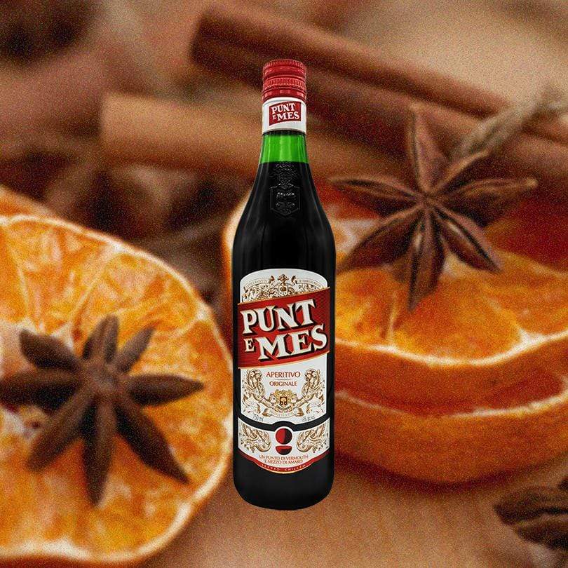 Bottle of Carpano Punt E Mes Sweet Vermouth over backdrop of sliced oranges and spices on a table.