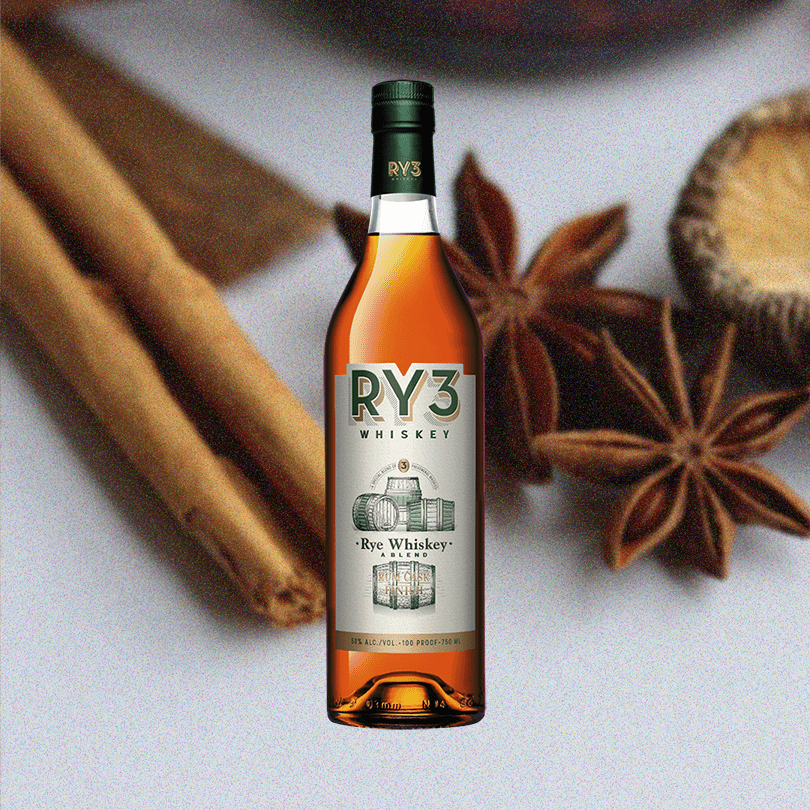 Bottle of RY3 Whiskey Rum Cask Finish over backdrop of cinnamon and similar spices on a table.