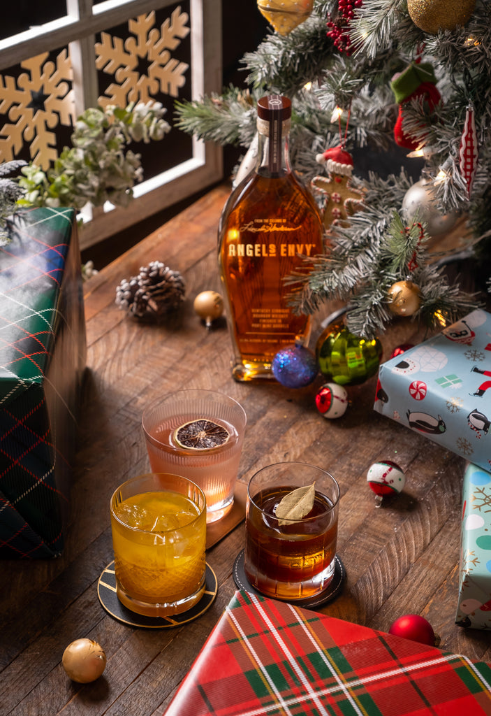 Bottle of Angel's Envy Bourbon, along with cocktails and presents, all placed under a Christmas tree.