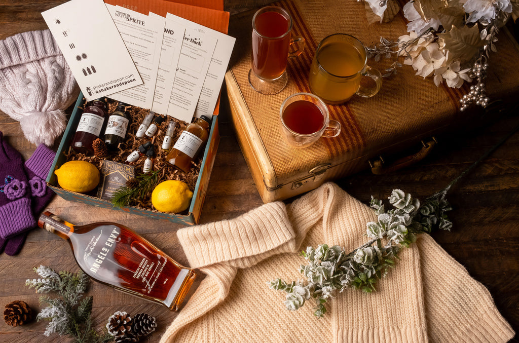 Bottle of Angel's Envy Bourbon along with Shaker and Spoon gift box, cocktails and an arrangement of winter decor.