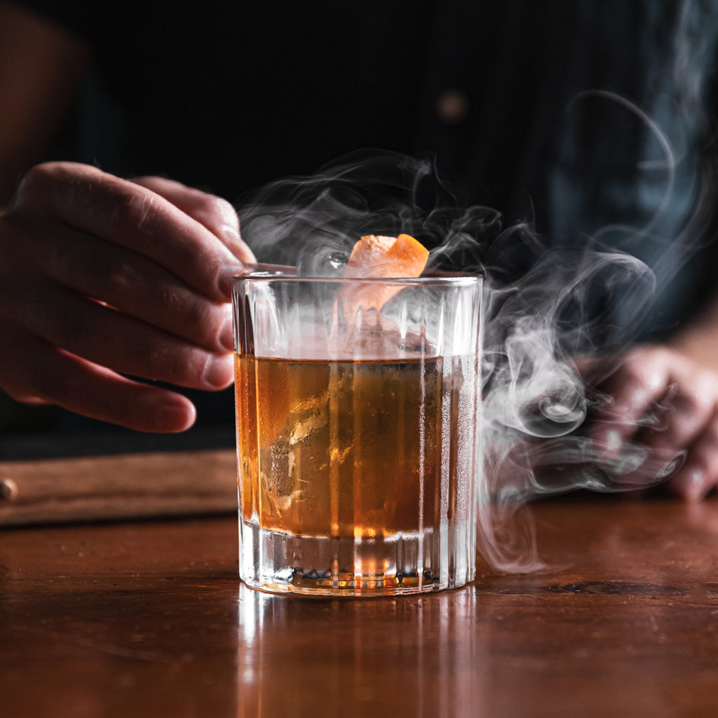Old fashion cocktail with smoke surrounding, with hand placing an orange garnish on top.