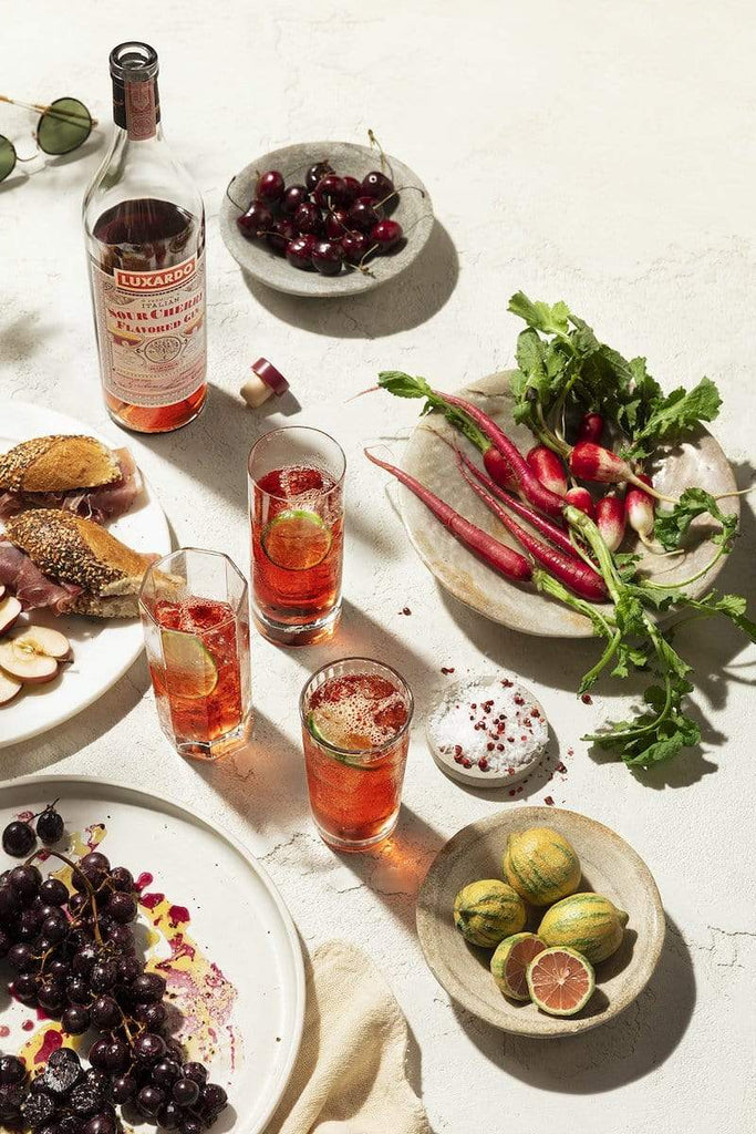 Table scape with Luxardo Sour Cherry Gin, cocktails, sandwiches, maraschino cherries, and root vegetables.