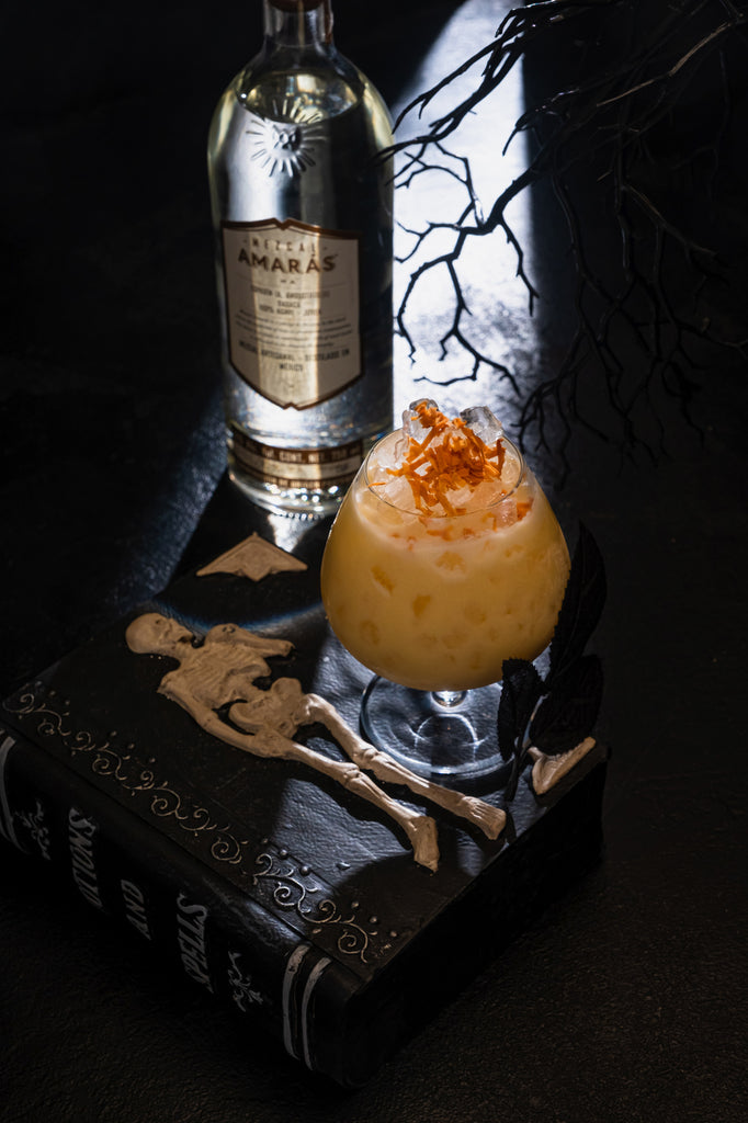 Bottle of Mezcal Amarás Espadin Joven next to a spooky cocktail and black book.