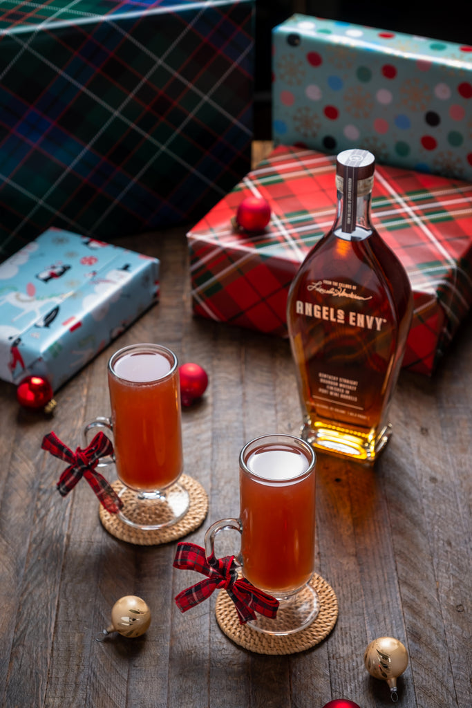 Bottle of Angel's Envy Bourbon along with festive looking cocktails and wrapped presents in background.