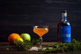 Bottle of Balcones Baby Blue Corn Whisky on a wooden table next to a minimal cocktail, lemon, apple and orange.