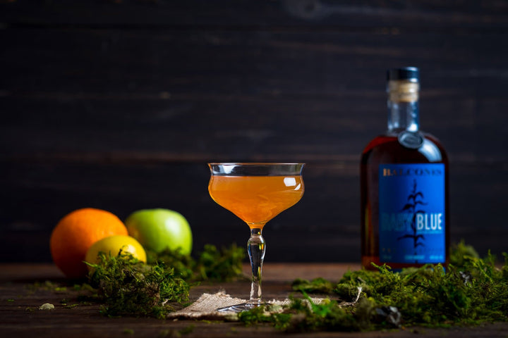 Bottle of Balcones Baby Blue Corn Whisky on a wooden table next to a minimal cocktail, lemon, apple and orange.