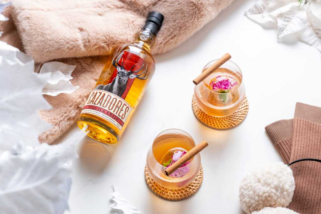 Bottle of Cazadores Añejo Tequila lying on table, next to plush blanket and two cocktails with cinnamon garnish.