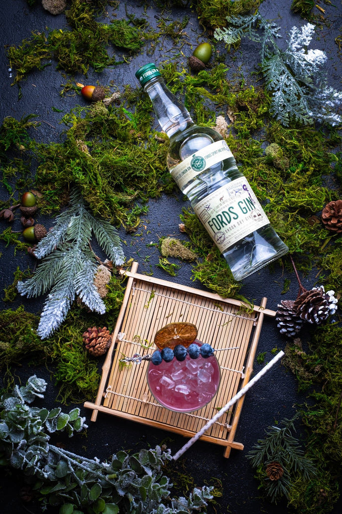 Bottle of Fords Gin on the ground surrounded by greenery next to a blueberry cocktail.