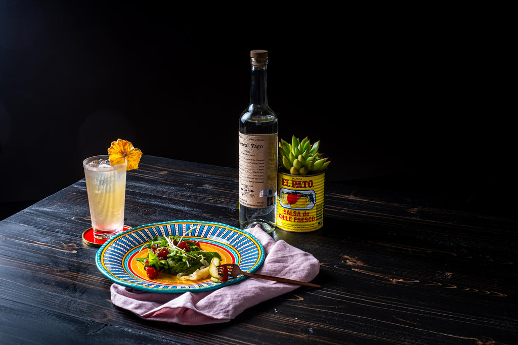 Bottle of Mezcal Vago Elote on a table in a dark room next to a plate, cocktail and plant.