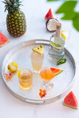 Bottle of Novo Fogo Silver Cachaça on a tray next to multiple cocktails and scattered fresh fruit.
