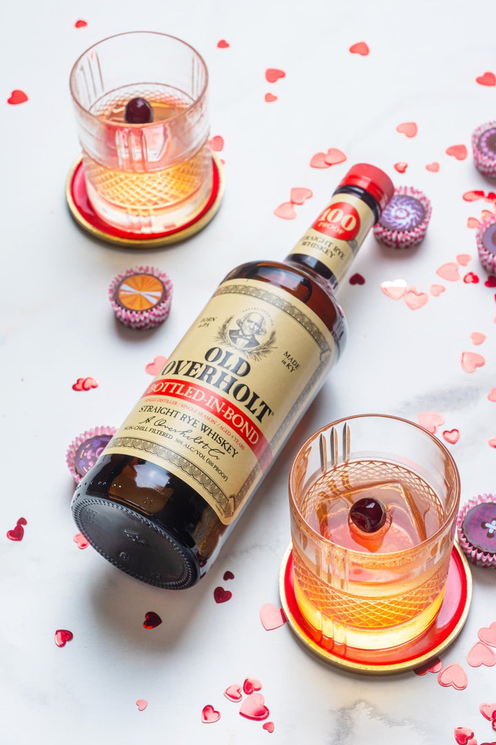 Bottle of Old Overholt Bonded Straight Rye Whiskey on a table next to two cocktails and scattered Valentine's Day decorations.