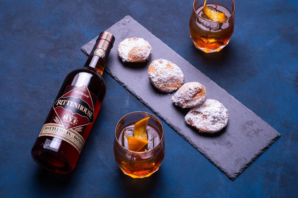 Bottle of Rittenhouse Straight Rye Whiskey on a blue carpet next to baked goods and two cocktails.