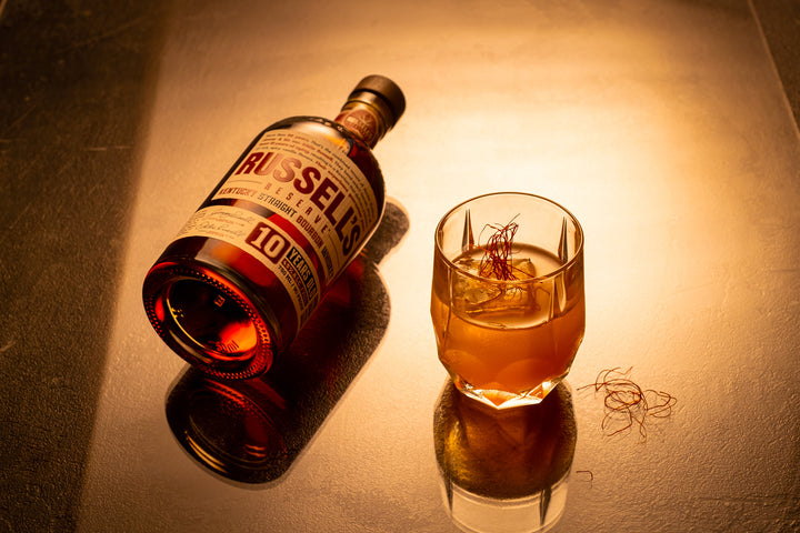 750ml bottle of Russell's Reserve 10 Year Bourbon in dark lighting on a table next to a cocktail.