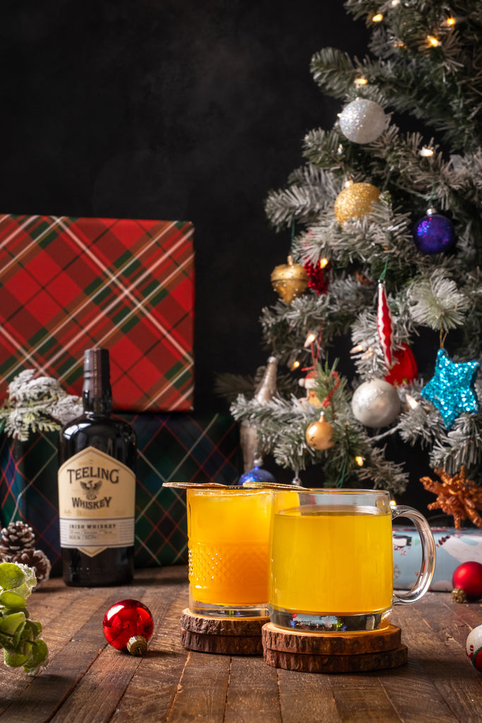 Bottle of Teeling Small Batch Irish Whiskey in background alongside Christmas tree and packages, and two cocktails out front sitting on wooden coasters.
