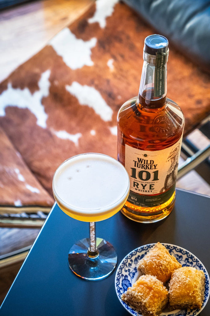 Bottle of Wild Turkey 101 Kentucky Straight Rye Whiskey on the corner of a table next to baked goods and a light cocktail.