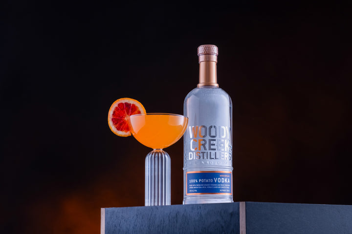 Bottle of Woody Creek Potato Vodka on a table next to an orange cocktail.