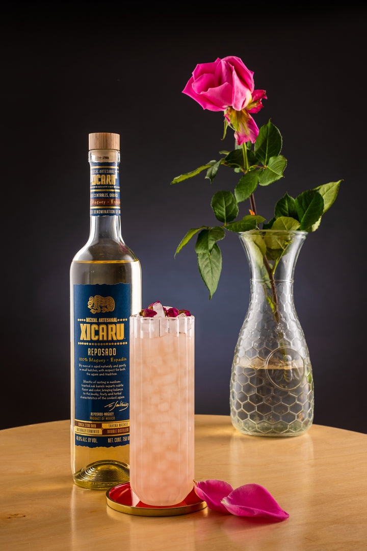 Bottle of Xicaru Mezcal Reposado on a table next to a pink rose in a vase and a dainty pink cocktail.