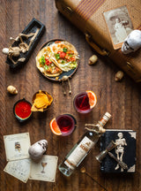 Bottle of Xicaru Silver Mezcal on a wooden floor next to trincits, tacos, and cocktails.