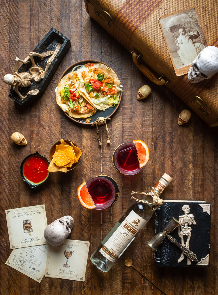 Bottle of Xicaru Silver Mezcal on a wooden floor next to trincits, tacos, and cocktails.